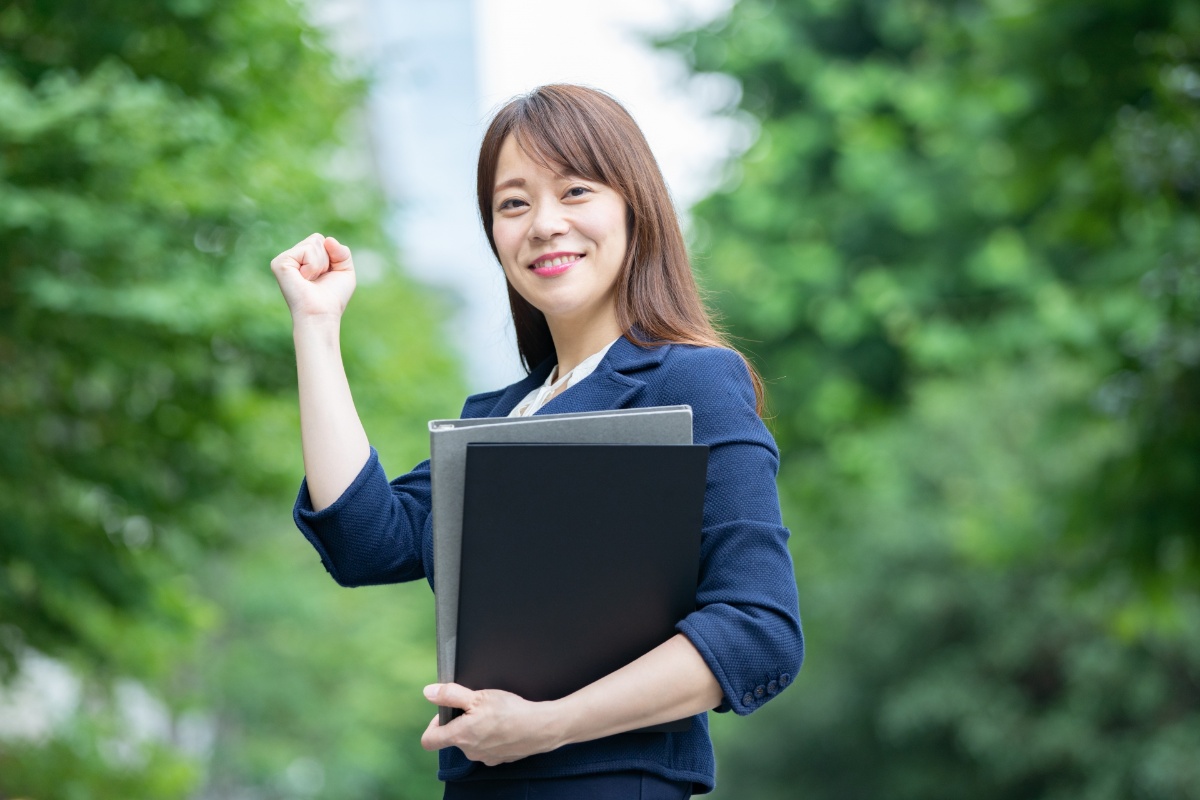 紺色のジャケットを着た若い日本人女性が、ノートパソコンを抱えながら、ガッツポーズで勝利の笑顔を浮かべている。背景には緑の樹木が広がる屋外の公園。成功と達成を表現したビジネスウーマンのポートレート